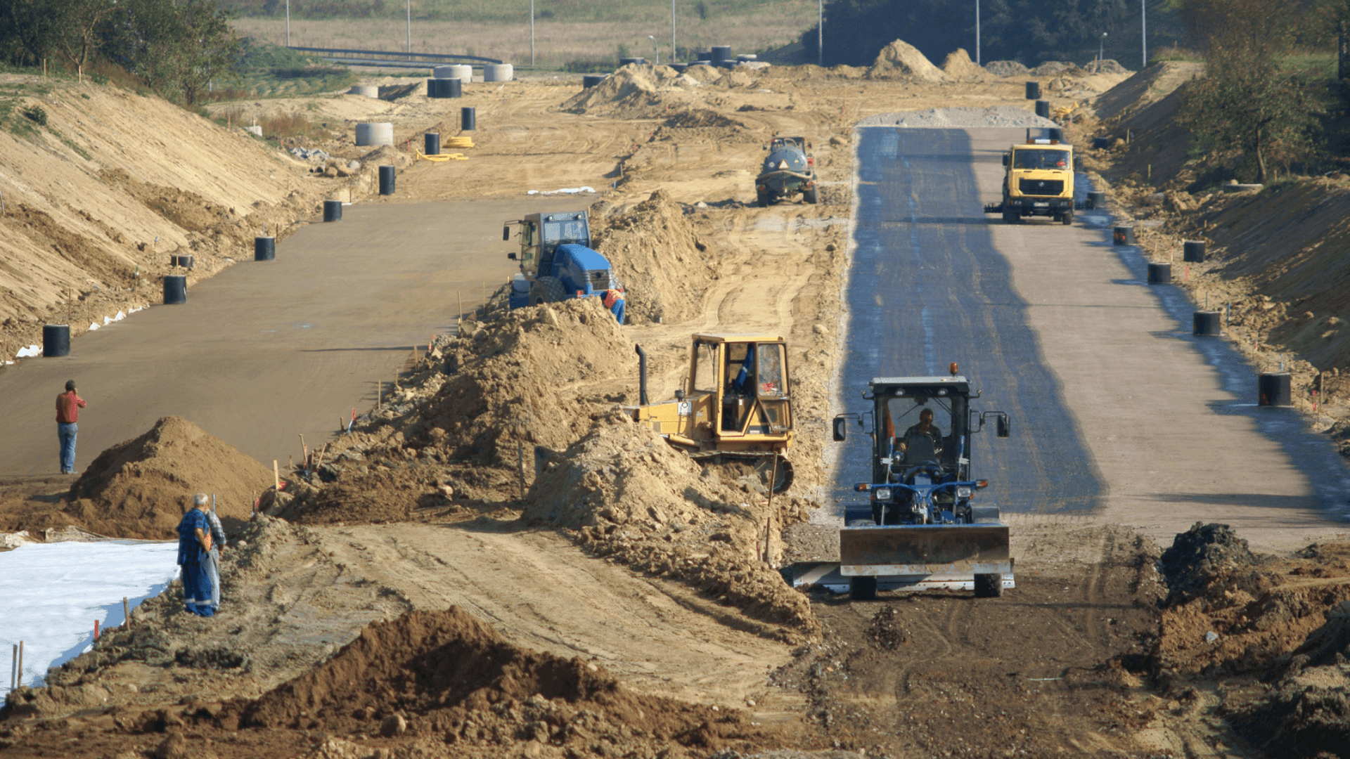 Construction site with heavy machinery working on a road, surrounded by dirt mounds and construction workers.