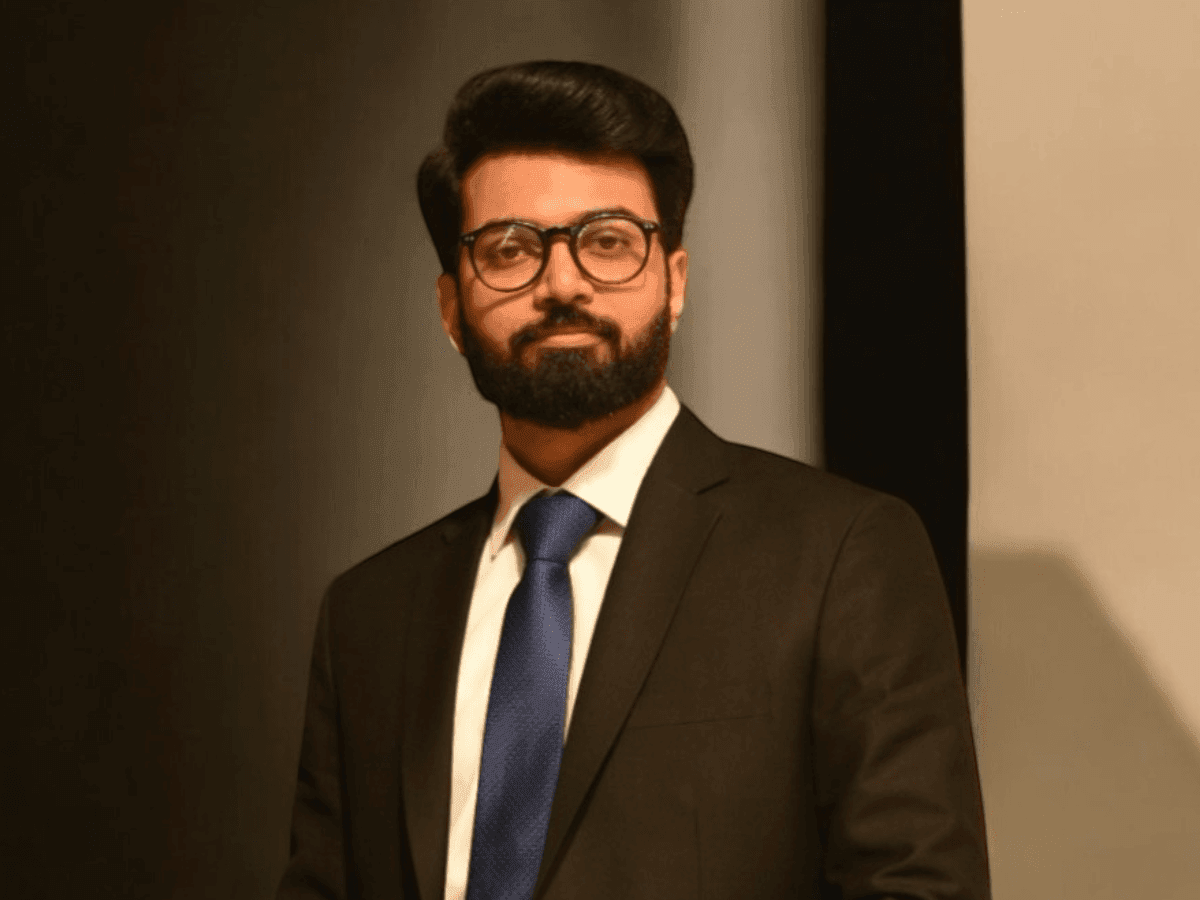 Man in a suit and tie standing against a dark background, wearing glasses and a beard.